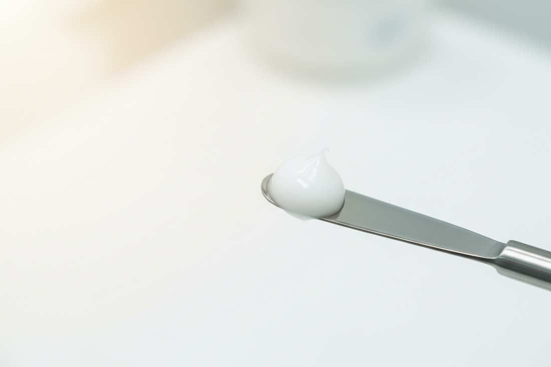Minimalist macro photography of a clinical white topical base on a stainless steel spatula, representing the precision of Mixwell Pharmacy's customized sports medicine